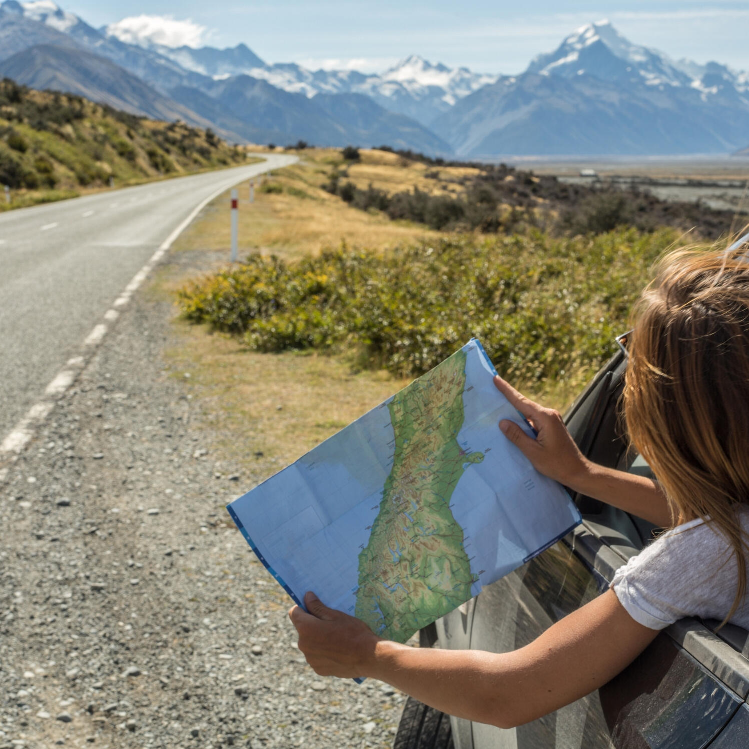 mountain-road-trip-map-travel-woman Female traveller holding a map out of a car window on a scenic mountain road trip, surrounded by rugged peaks and open highway adventure.